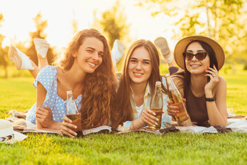 Three smiling girlfriends, girlfriends drink cold drinks and chat, lying on a green lawn outdoors on a sunny day in the park