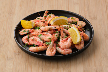 Boiled shrimp garnished with lemon and parsley in a dark plate on a light wooden background. View from above.