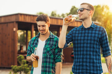 Fototapeta premium Two young male friends walking in the park in summer on a sunny day, talking and drinking beer drinks
