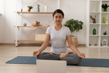 Fototapeta na wymiar Healthy Indian woman sit on mat at home practice yoga with online class or training on laptop. Calm young mixed race female in sportswear exercise medicate with video webcam computer lesson.