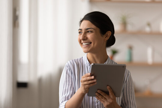 Smiling young Indian woman use modern tablet gadget look in distance dreaming visualizing success perspective. Happy mixed race female browse internet or talk on video call on pad device.