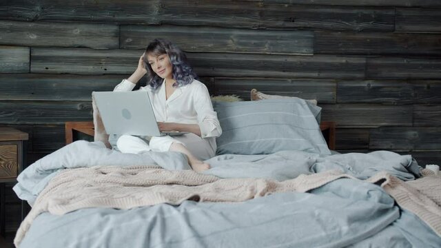 Slow motion of cheerful young woman using laptop in bed and smiling enjoying looking at screen and internet content. Youth and devices concept.