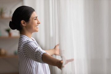 Overjoyed young Indian woman open curtains look in window distance meet welcome new day at home....