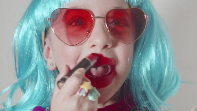 Adorable little child girl wearing sunglasses and a blue wig paints her lips with lipstick, plays learn makeup in front of the small mirror. Imitates adults making experiments with cosmetics, eyeglass