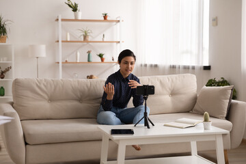 Obraz premium Young Indian woman sit on couch at home record video blog or live broadcast on smartphone. Millennial confident mixed race female coach talk speak on webcam virtual online call on cellphone.