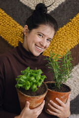 Happy young woman holding fresh green basil plant and rosemary plant in clay pots  and smiling on background of rustic room. Repotting and cultivating aromatic herbs at home. Horticulture