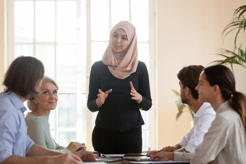 Smiling Asian muslim team leader talking to diverse employees at briefing, businesswoman wearing...