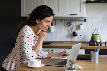 Confident smiling businesswoman using laptop in kitchen, working on project online at home, focused young woman looking at computer screen, writing taking notes, student watching webinar, studying