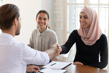 Smiling Asian muslim businesswoman wearing hijab shaking businessman hand at meeting, celebrating...