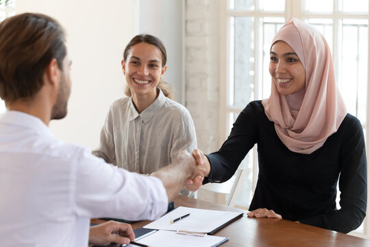 Smiling Asian muslim businesswoman wearing hijab shaking businessman hand at meeting, celebrating contract signing, diverse business partners making deal, group negotiations in boardroom