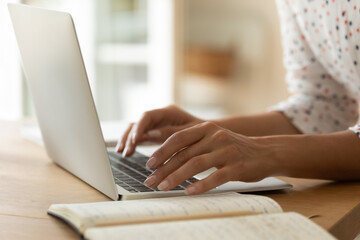 Close up young woman working online on laptop, chatting or browsing device apps, sitting at wooden table, cropped of female hands typing on computer keyboard, writing message in social networks