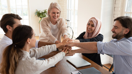 Excited smiling multiethnic employees team stacking hands at corporate meeting, sitting at table in boardroom, happy business people celebrating shared success, motivated by good teamwork result