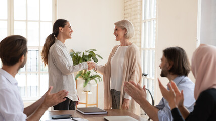 Mature executive shaking successful businesswoman hand at corporate meeting, middle aged team leader greeting new employee, young intern or thanking for good work result, colleagues applauding