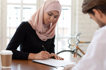 Asian muslim businesswoman wearing hijab signing job contract or agreement with business partner at meeting, saleswoman putting signature on legal documents, making investment or insurance deal - Powered by Adobe