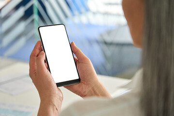 Female hands holding using cell mobile smartphone with empty blank white screen for modern application advertising at desk. Business technologies concept. Over shoulder view.