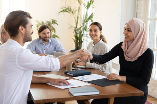 Smiling Asian muslim businesswoman shaking hand of businessman making agreement at meeting in boardroom, group negotiation, diverse business partners handshaking, celebrating contract signing