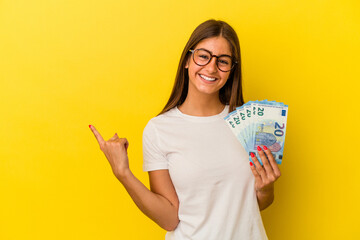 Young caucasian woman holding bills isolated on yellow background smiling and pointing aside,...