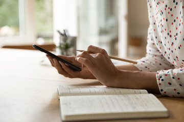 Close up cropped of woman holding smartphone and pencil, taking notes in notebook, important...