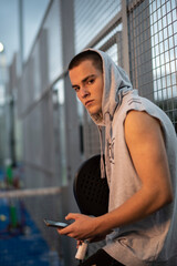 Young man with urban style takes a break with the phone trains after playing paddle tennis on an outdoor court