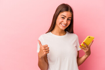 Young caucasian woman holding a mobile phone isolated on pink background smiling and raising thumb...