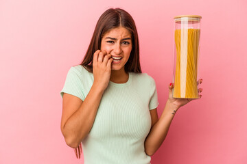 Young caucasian woman holding a pasta jar isolated on pink background biting fingernails, nervous and very anxious.