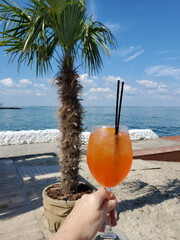 POV female hand holding wine glass of luxury cocktail Aperol Spritz on tropical beach with amazing ocean view. Summer vacation party drink. Vertical shot