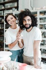 Happy couple of african american guy and asian girl are standing next to each other in a maker's shop and smiling