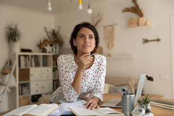 Dreamy thoughtful woman pondering research project, studying at home, standing at table with books and laptop, motivated young female student taking notes, writing essay, homework appointment