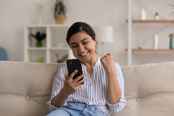 Overjoyed young Indian woman look at smartphone screen triumph celebrate win or victory online. Happy excited mixed race female feel euphoric with good promotion or hiring news on cellphone.