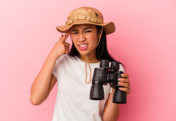 Young mixed race explorer woman holding binoculars isolated on pink background showing a disappointment gesture with forefinger.