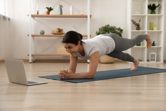 Happy millennial Indian female in sportswear on mat at home practice yoga stretching with online lesson or class on computer. Smiling ethnic woman train do workout watch video session on laptop.