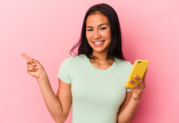 Young Venezuelan woman holding a mobile phone isolated on pink background smiling and pointing aside, showing something at blank space.