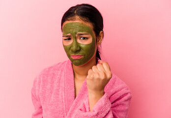 Young Venezuelan woman wearing a bathrobe and facial mask isolated on pink background showing fist to camera, aggressive facial expression.