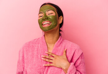 Young Venezuelan woman wearing a bathrobe and facial mask isolated on pink background laughs out loudly keeping hand on chest.