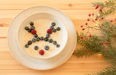 Obraz na płótnie Canvas Kid food decoration, funny christmas deer made of ripe berries in a plate on a decorated wooden table. Creative idea of kid's dishes for a winter holiday. Milk oatmeal with blueberry, raspberry decor.
