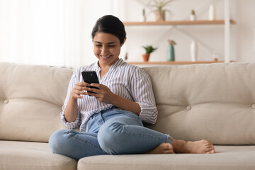 Obraz premium Happy millennial Indian woman sit relax on sofa at home talk speak on video call. Smiling young mixed race female rest on couch have webcam online digital communication on smartphone.