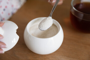 Close up cropped of woman putting spoon of granulated sugar in black tea from white porcelain bowl,...