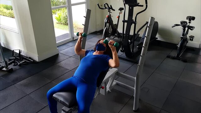 74 year old woman with mask doing incline bench press with dumbbells.