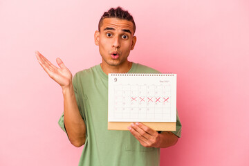 Young venezuelan man holding a calendar isolated on pink background surprised and shocked.