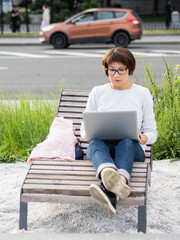 Woman sits with laptop on urban park bench. Freelancer at work. Student learns remotely from outdoors. Modern lifestyle.