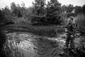 A fisherman dressed in camouflage fishing in the river. Black and white photo.