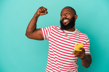 African american man holding a mobile phone isolated on blue background raising fist after a...