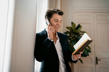 Fototapeta premium An adult male lawyer works in his office in a tablet and calls. Makes a design project remotely writes down new ideas in the diary. Dressed in a modern business suit.
