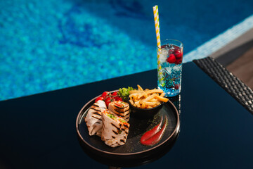 Chicken in a tortilla with cherry tomatoes and french fries placed by the pool next to a summer cocktail with strawberries. Pool party