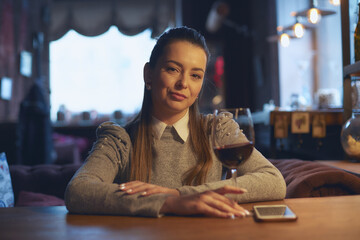 A beautiful young woman sits at a wooden table in the premises of a wine bar and drinks wine from a glass.