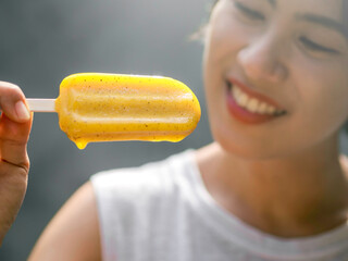 Close up yellow popsicle in beautiful happy Asian woman’s hand wearing casual white sleeveless...