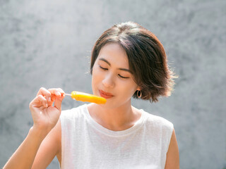 Woman eating popsicles. Happy beautiful Asian woman wearing casual white sleeveless shirt holding yellow popsicle, outdoors. Smiling female enjoying ice lolly in summer.