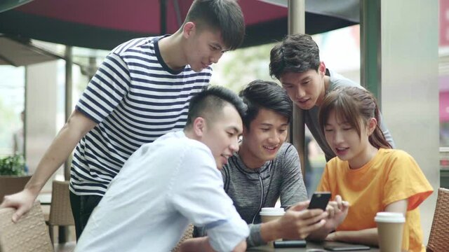 group of young asian adults four men and a woman using looking at mobile phone together outdoors in a coffee shop