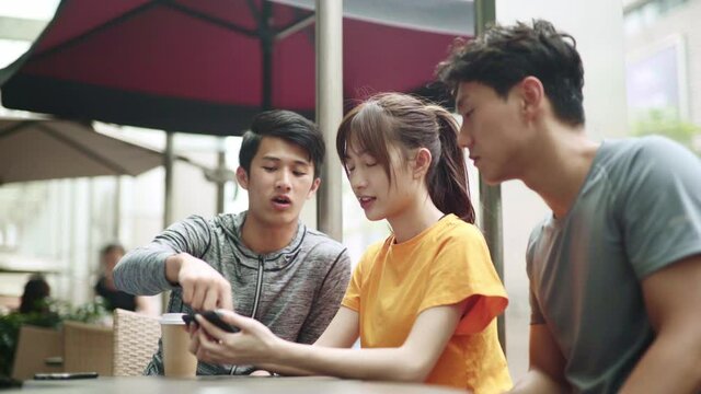 three asian young adults two men and a woman sitting in outdoor coffee shop looking at cellphone together