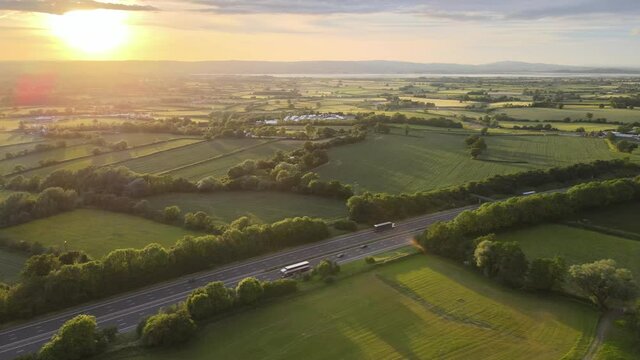 Aerial View Of Vehicles Driving At M5 Motorway Passing By Green Fields At Dursley Town At Sunrise In England, UK.